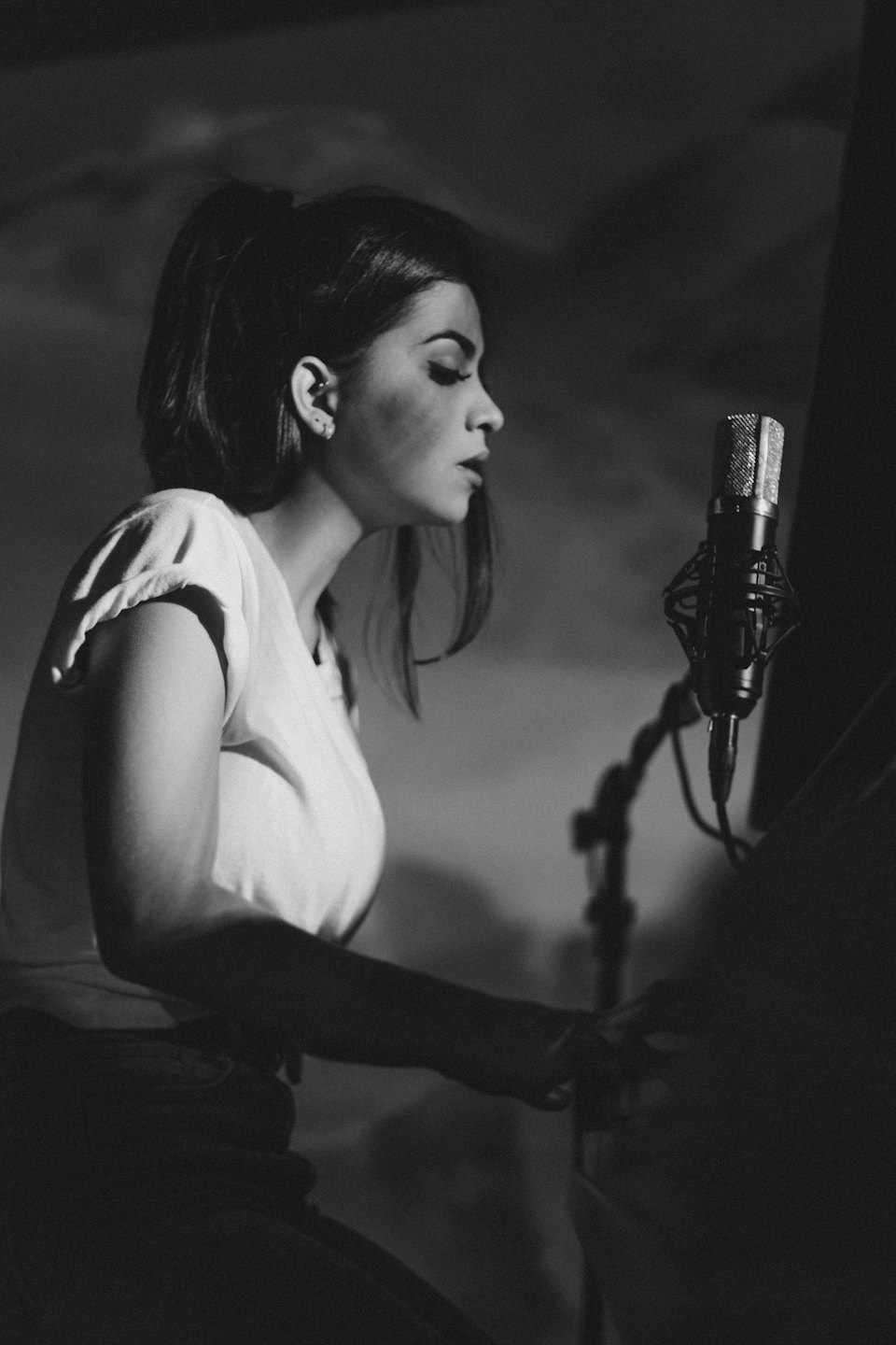 Woman singing into a studio microphone with hands near keyboard.
